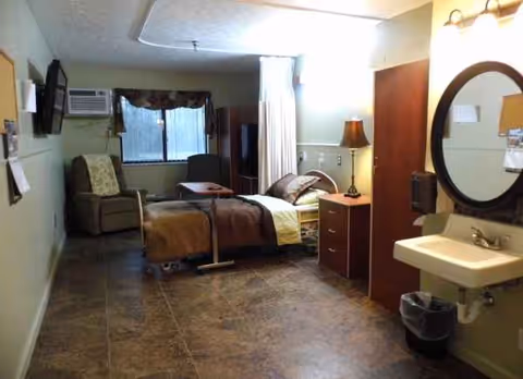 Senior living facility bedroom with bed, chair, dresser, sink, and round mirror.