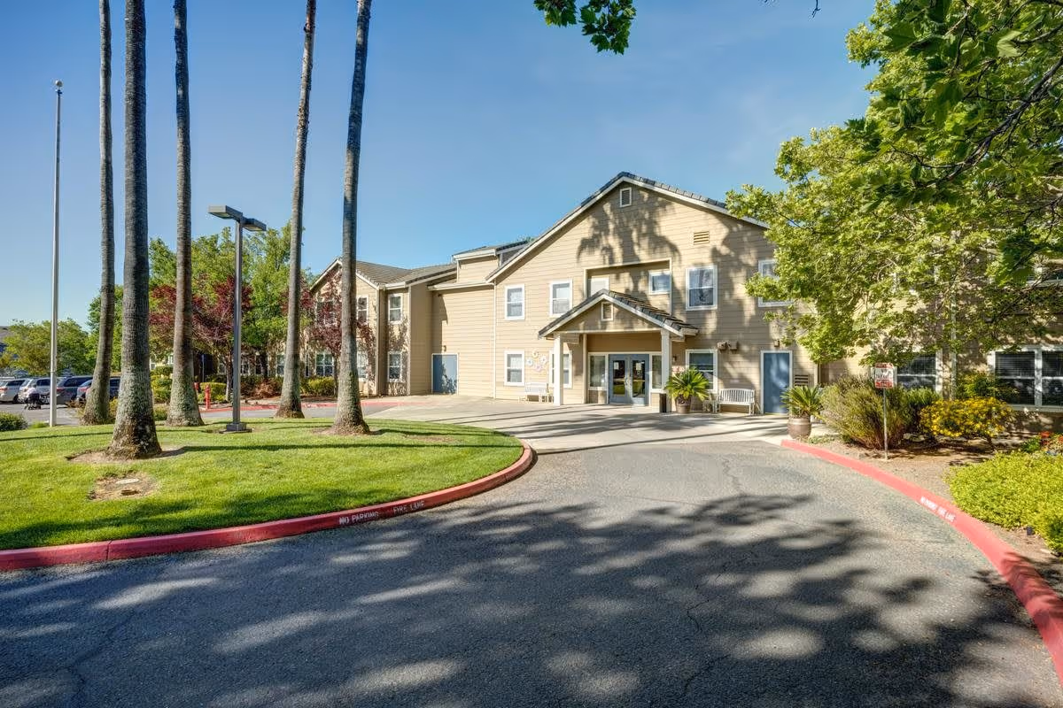 Exterior view of a senior living facility building with beige siding, multiple windows, and a covered entrance. The driveway curves in front of the entrance, bordered by a red-painted curb. Tall palm trees and other green trees and shrubs surround the building under a clear blue sky.
