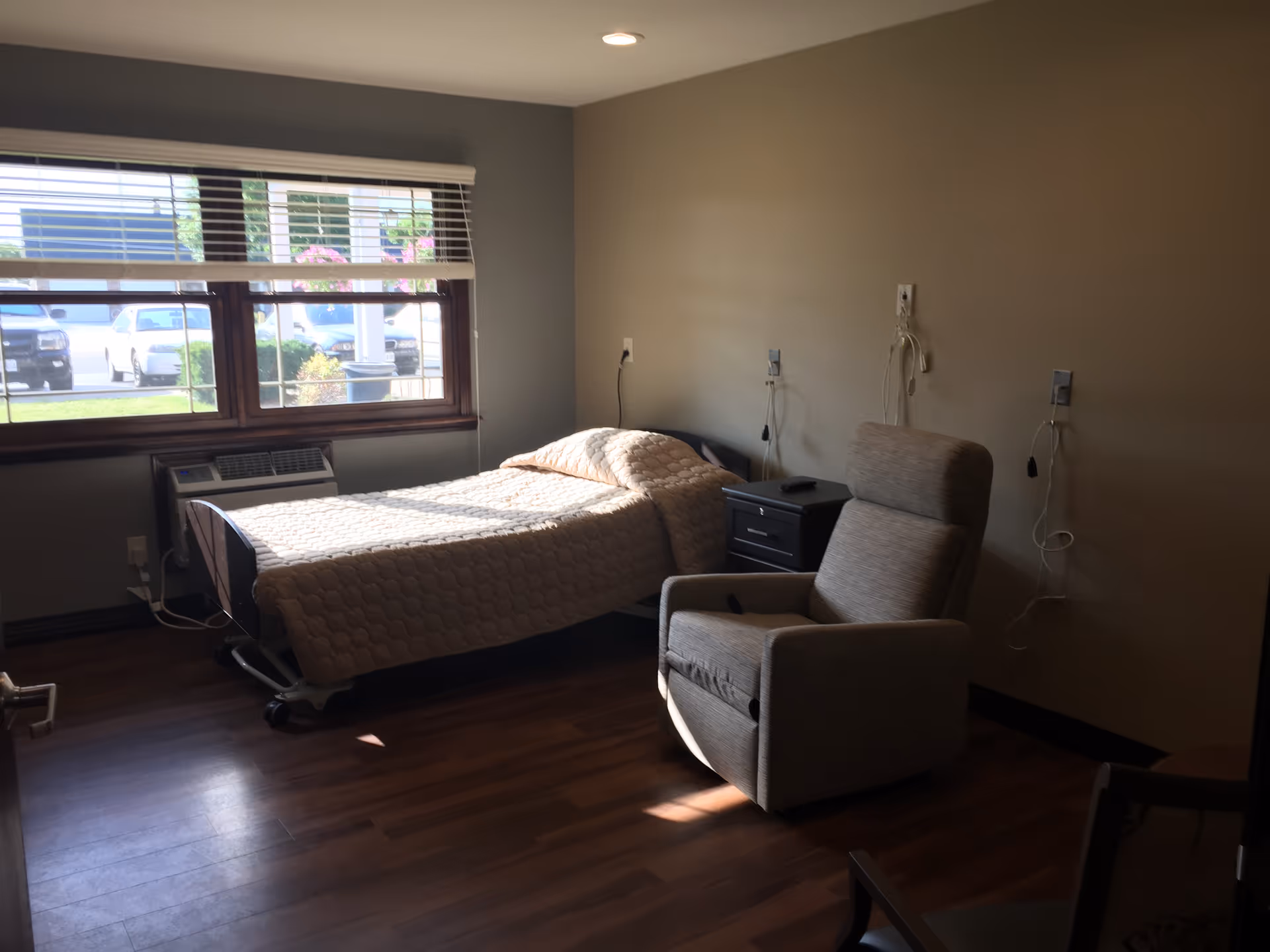 A simple bedroom in a senior living facility with a single bed covered in a beige quilt, a gray recliner chair, a small black nightstand, and a large window with blinds letting in natural light. The room has wooden flooring and beige walls with medical call buttons and cords mounted on the wall.