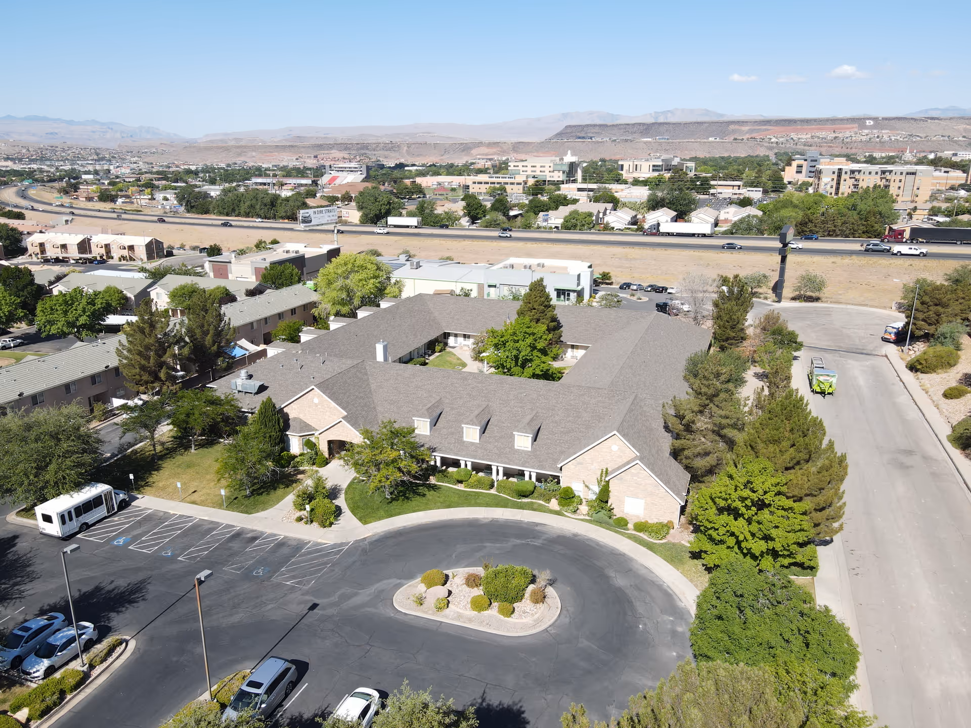 Aerial view of a single-story assisted living building with a central courtyard, surrounding trees, parking lot, and nearby highway.