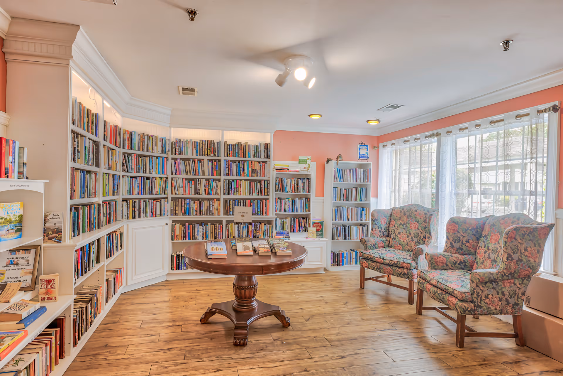 A cozy library room with wooden floors, white built-in bookshelves filled with books, a round wooden table in the center with books displayed on it, and two floral upholstered armchairs near large windows with sheer white curtains allowing natural light to fill the space.