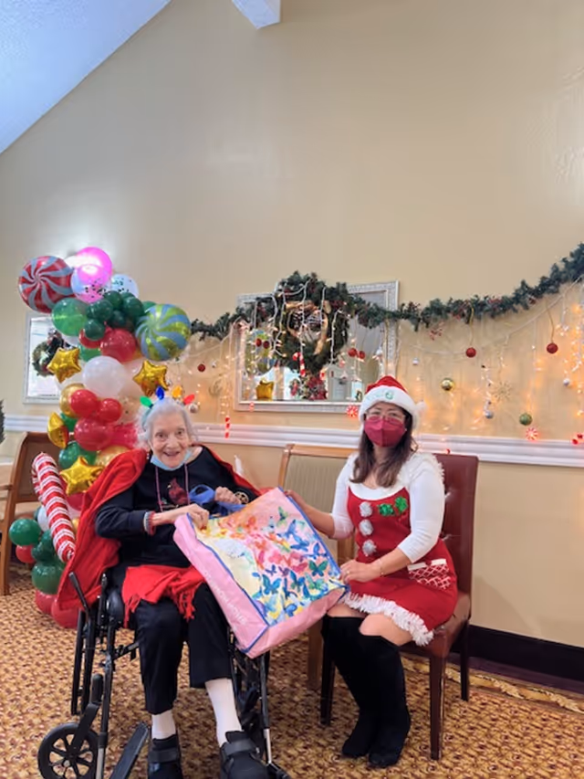 An elderly woman in a wheelchair and a woman dressed in a festive holiday outfit with a Santa hat and red mask sit together in a decorated room. The elderly woman is holding a colorful gift bag. The background features Christmas decorations including garlands, lights, ornaments, and balloon arrangements shaped like candy and stars.