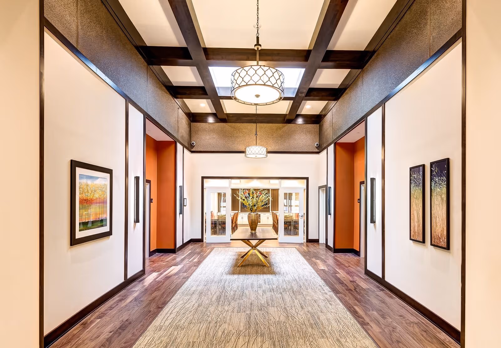 A bright, elegant hallway with wooden flooring and a patterned rug leading to a dining area. The hallway features a coffered ceiling with two hanging light fixtures, framed artwork on the walls, and a central table with a large vase of flowers.