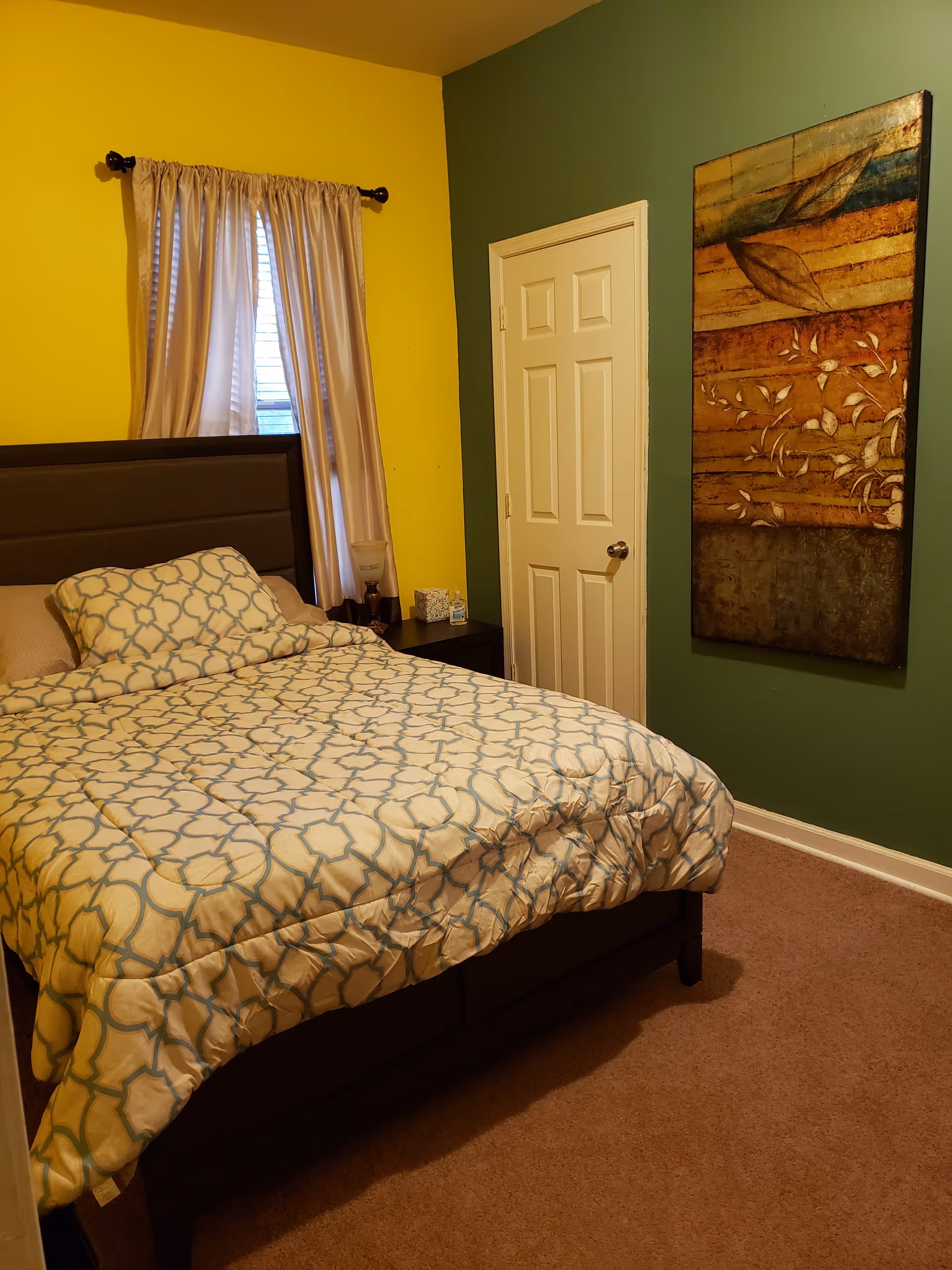 A bedroom with a bed covered in a patterned comforter and matching pillow. The walls are painted yellow and green. There is a window with beige curtains above a nightstand holding a lamp and a box of tissues. A door is visible on the green wall, and a large decorative painting hangs next to it.