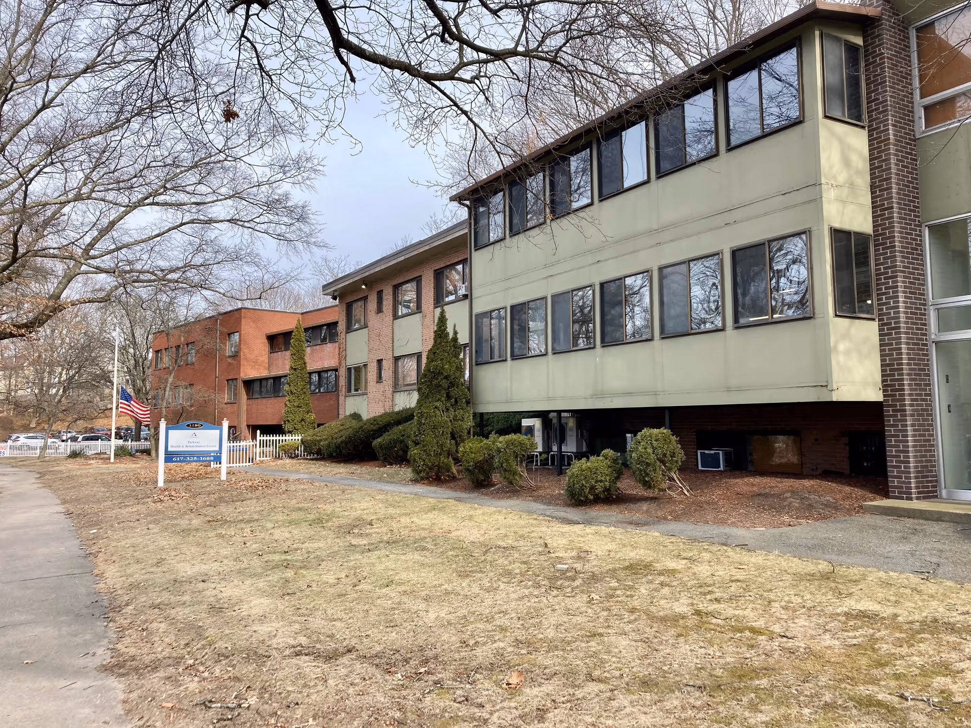 Exterior view of Parkway Health & Rehabilitation Center, a multi-story building with large windows. The building is surrounded by leafless trees and some bushes, with a sidewalk and a grassy area in the foreground. An American flag is visible near a sign in front of the building.