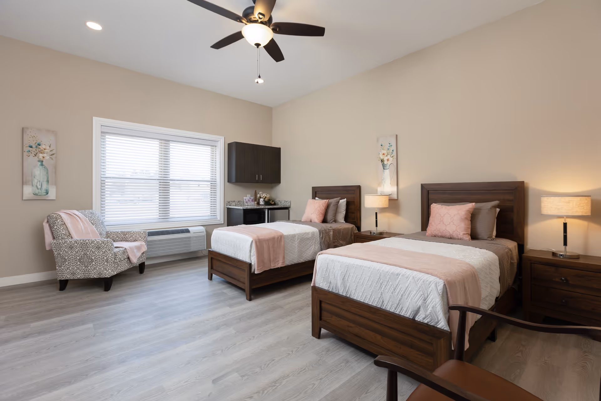 A bright and spacious bedroom with two single wooden beds, each with white and pink bedding and pillows. There are two wooden nightstands with lamps on either side of the beds. A patterned armchair with a pink throw is placed near a large window with white blinds. Above the beds, there are floral paintings on the beige walls. A ceiling fan with a light is mounted on the ceiling. A small cabinet with a countertop and a microwave is positioned between the window and the beds.