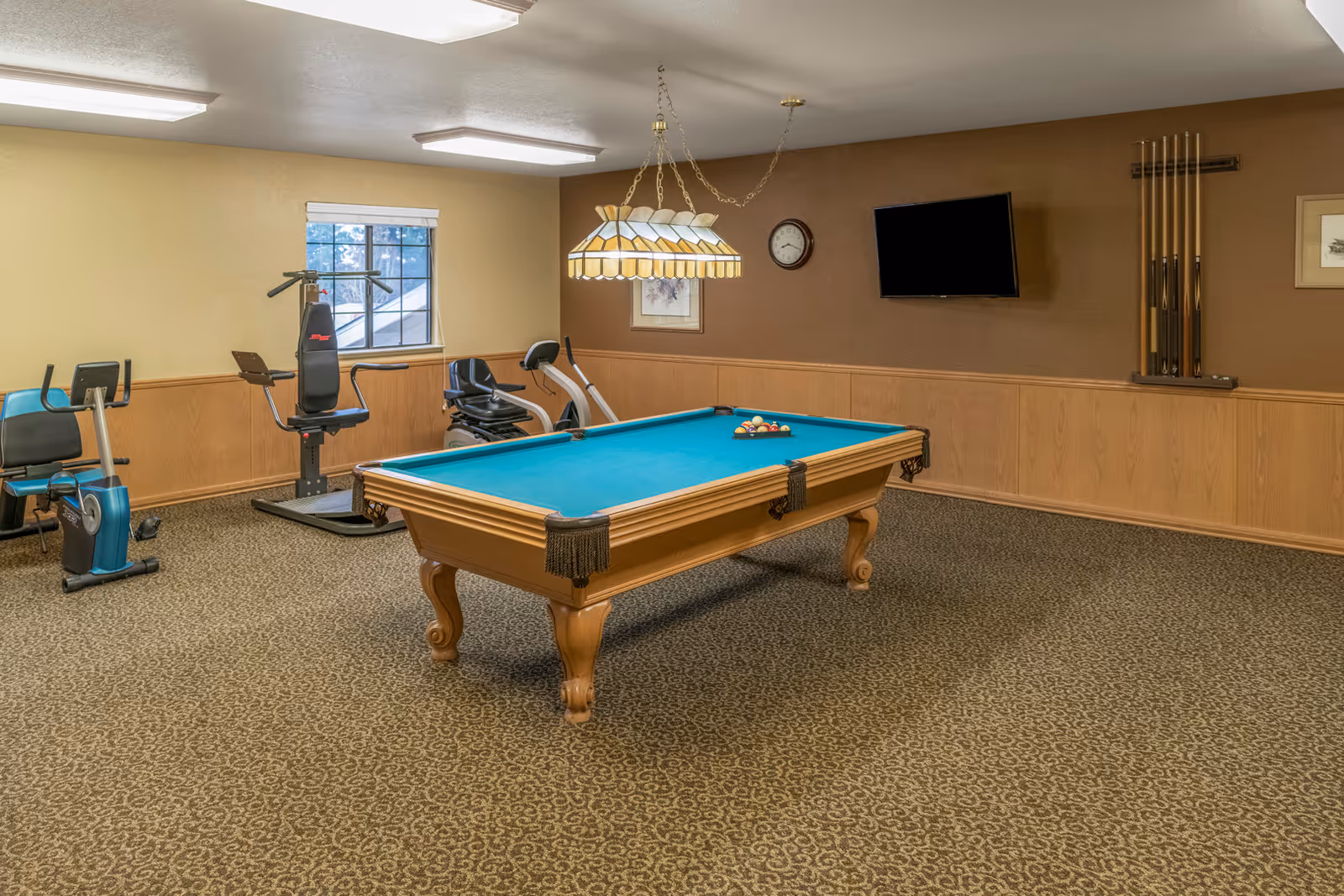 A recreational room featuring a pool table with a green felt surface and a hanging stained glass light fixture above it. In the background, there are exercise machines including a stationary bike and a multi-exercise machine. The room has beige walls with wood paneling, a wall-mounted TV, a clock, and a rack holding pool cues. The floor is carpeted with a patterned design.