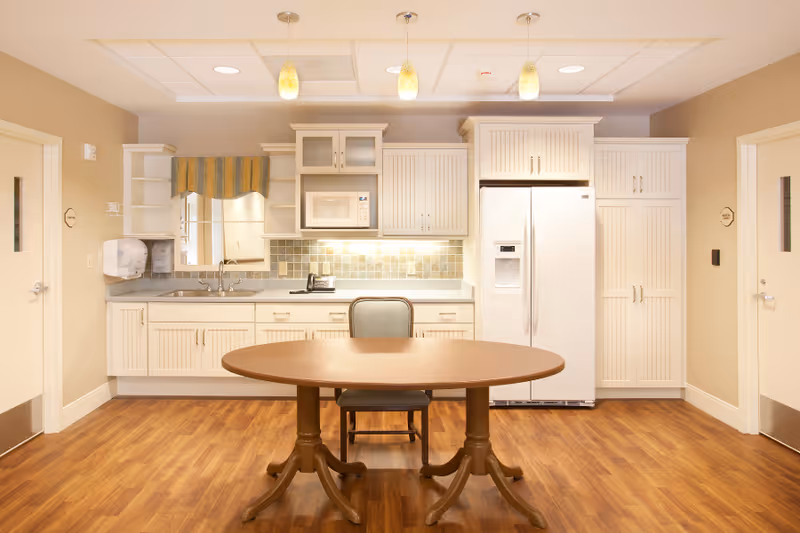Communal kitchen with white cabinetry, refrigerator, microwave, sink, and a round wooden table with a chair in the center.