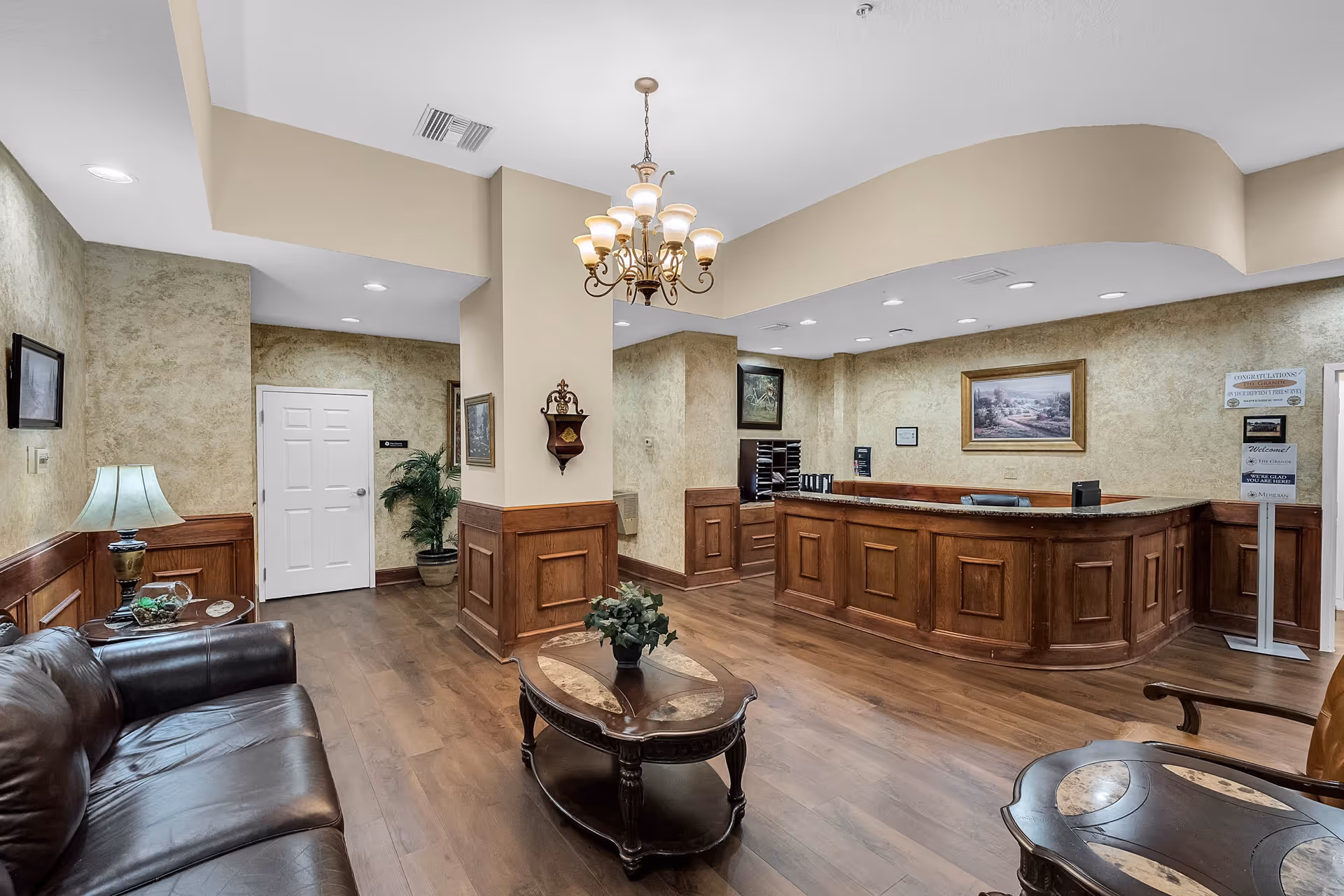 A reception area in a senior living facility with a wooden front desk, a chandelier hanging from the ceiling, a leather sofa, wooden coffee tables with decorative plants, framed artwork on the walls, and a door leading to another room.