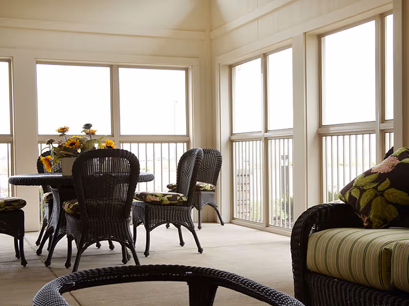 A sunlit indoor seating area with wicker chairs and a round table. The chairs have floral and striped cushions. Large windows with white frames let in ample natural light, creating a bright and airy atmosphere.