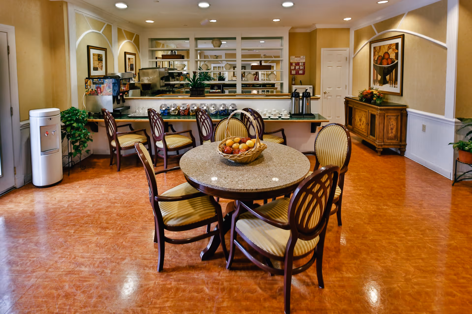 A cozy dining area with a round table surrounded by six cushioned chairs. A basket of fruit is placed on the table. In the background, there is a counter with coffee dispensers, cups, and a beverage dispenser. The room has warm lighting, wooden flooring, plants, and framed artwork on the walls.
