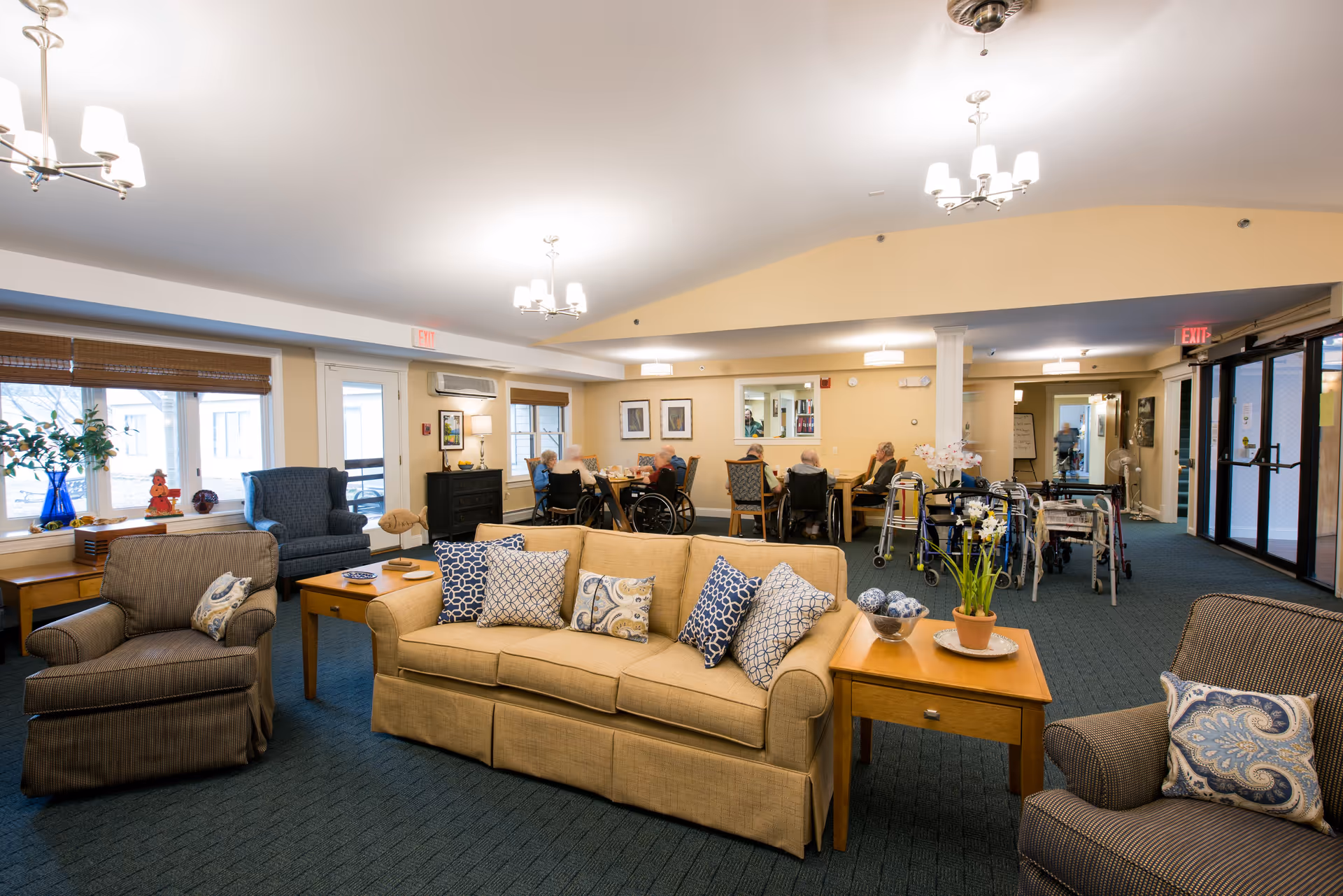 Spacious senior living common room with sofas and armchairs in the foreground and residents sitting at tables in the background.