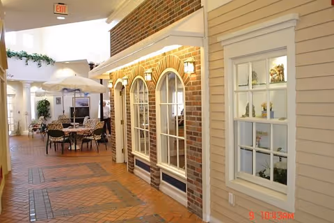 Indoor hallway area designed to resemble an outdoor street with brick and siding walls, arched windows, and a tiled floor. There are tables and chairs with umbrellas, plants, and a person standing in the background.