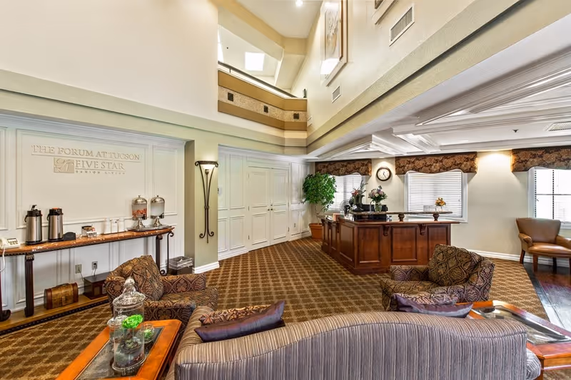 A spacious and well-lit senior living facility common area with patterned carpet, comfortable upholstered chairs and sofas, a wooden reception desk with flowers and a clock on the wall, large windows with valances, and a refreshment table with coffee dispensers and water containers. The walls are light-colored and the ceiling has recessed lighting.