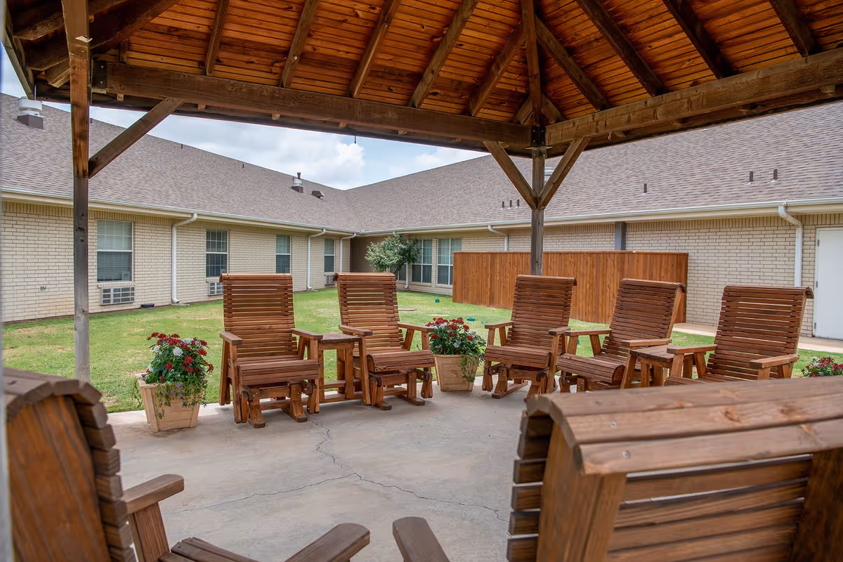 Outdoor covered seating area with wooden rocking chairs arranged in a circle on a concrete floor, surrounded by potted flowers and a grassy courtyard with a beige brick building in the background under a partly cloudy sky.