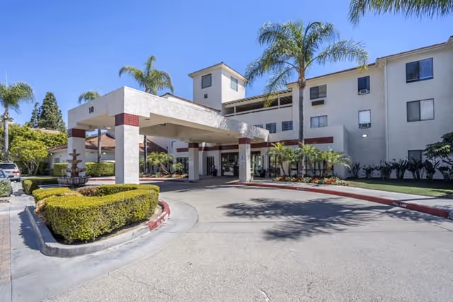Exterior view of Brookdale Irvine senior living facility showing the main entrance with a covered drop-off area, surrounded by palm trees and neatly trimmed bushes under a clear blue sky.