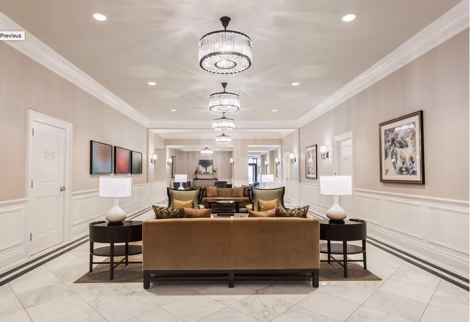 A spacious and elegant common area with beige walls, white wainscoting, and marble tile flooring. The room features multiple brown and green cushioned sofas arranged around dark wood coffee tables and side tables with white lamps. Crystal chandeliers hang from the ceiling, and framed artwork decorates the walls.