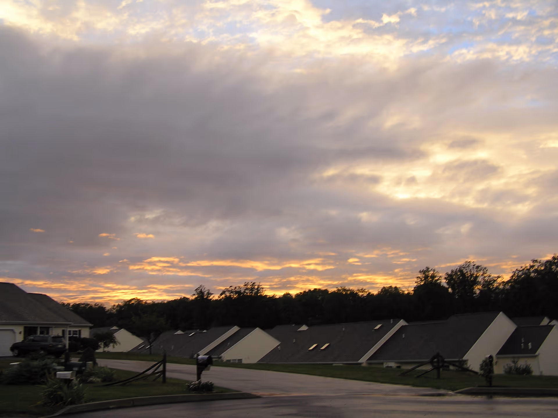 View of a residential street at sunset with a row of single-story houses with sloped roofs, trees in the background, and a cloudy sky with orange and yellow hues from the setting sun.