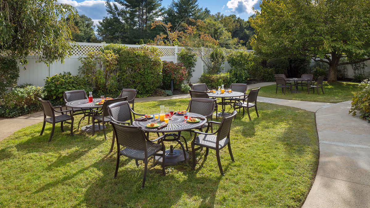 Outdoor garden area with several round metal tables and chairs arranged on a grassy lawn. Each table has plates with food and glasses of juice. The area is surrounded by green bushes, trees, and a white fence under a partly cloudy sky.