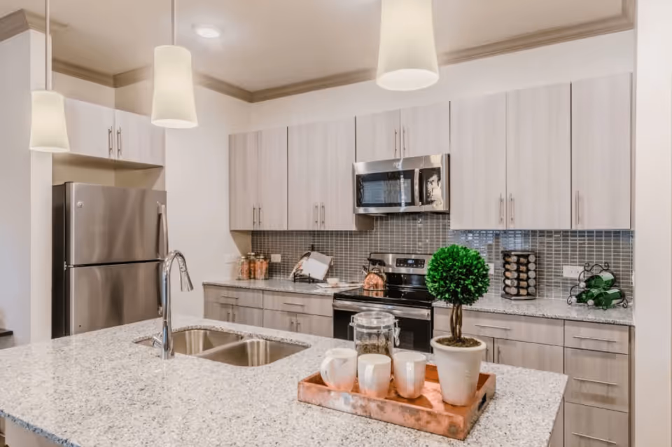 Modern kitchen with a granite island, stainless steel refrigerator and oven, pendant lights, and a decorative tray with a potted plant and mugs.