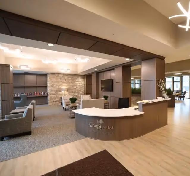 Reception area of The Woodlands of DeWitt featuring a curved wooden front desk with a white countertop, seating area with armchairs and sofas, a stone accent wall with a lamp, built-in bookshelves, and large windows letting in natural light.