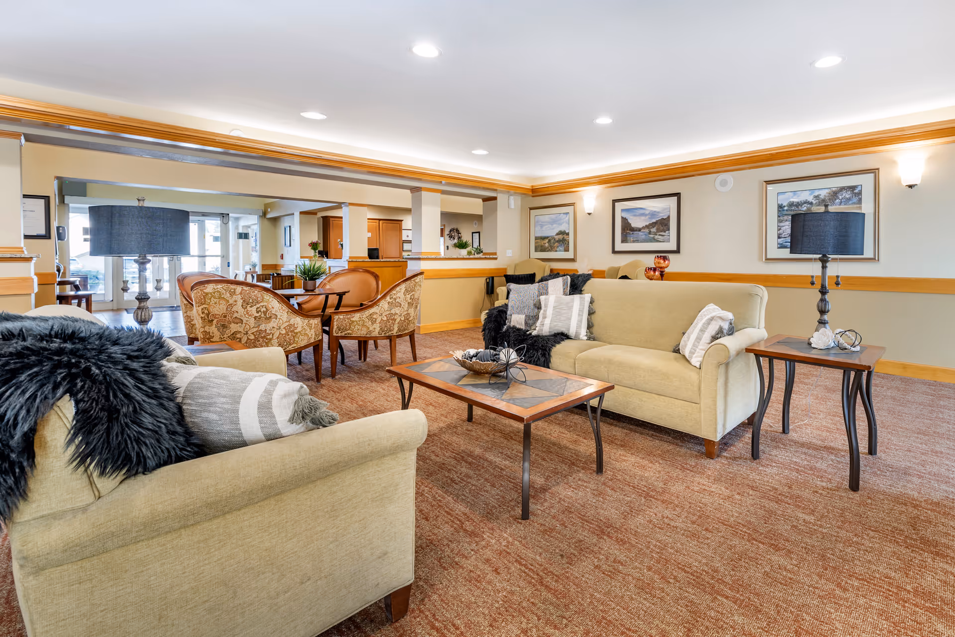 Bright, spacious senior living room lounge with sofas, armchairs, coffee tables, lamps, and framed artwork on the walls.