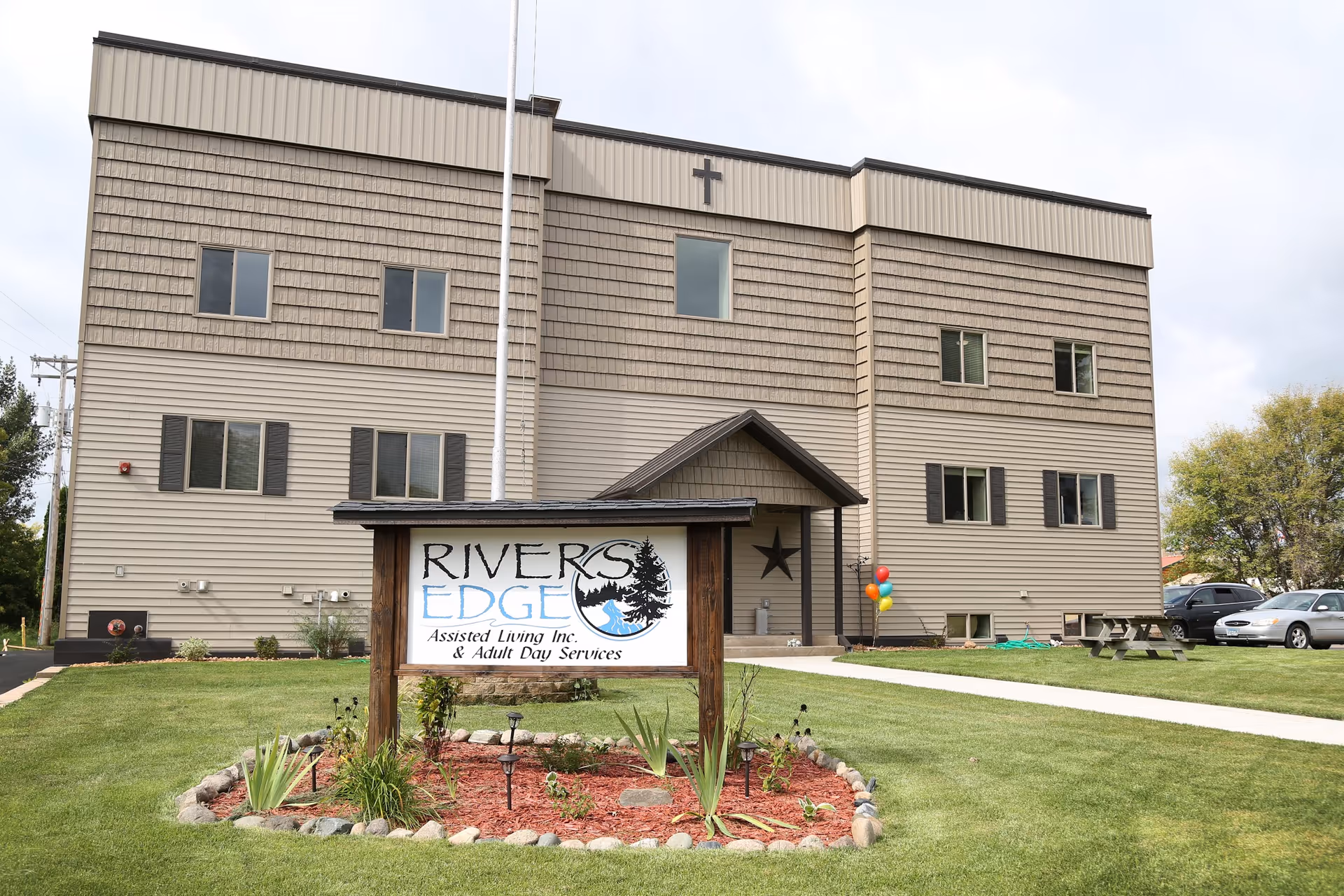 Exterior view of a three-story assisted living facility building with beige siding and a cross mounted near the roof. In front of the building is a landscaped area with a wooden sign that reads 'Rivers Edge Assisted Living Inc. & Adult Day Services'. There are a few parked cars and a picnic table on the lawn to the right.
