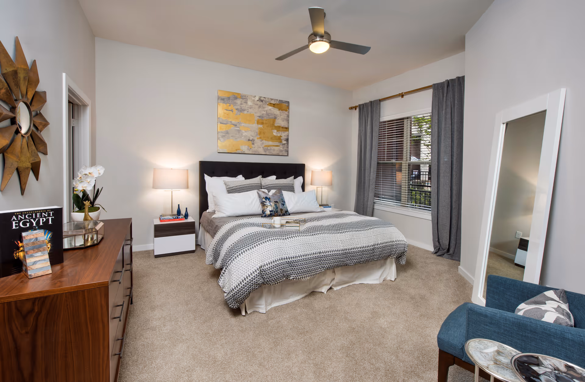 A well-lit bedroom featuring a large bed with patterned bedding and multiple pillows, flanked by two bedside tables with lamps. A wooden dresser with decorative items and a book titled 'Ancient Egypt' is on the left side. A large window with gray curtains is on the right wall, and a full-length mirror leans against the wall near a blue armchair with a patterned pillow. A ceiling fan with a light is mounted above.