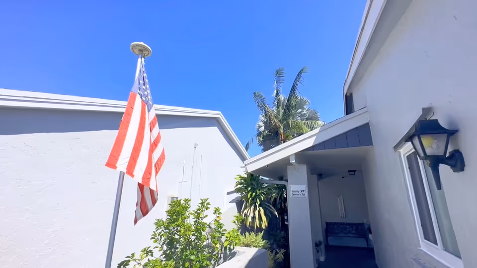 Outdoor view of a senior living facility with white buildings, an American flag on a flagpole, green plants, and a clear blue sky.