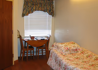 A small bedroom with a single bed covered in floral bedding, a wooden table with two chairs near a window with closed blinds and a floral valance, and a wooden door on the left side.