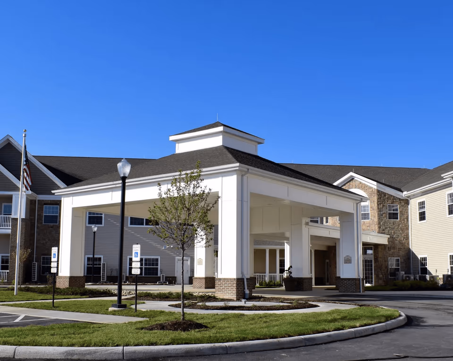 Exterior view of Danbury Huber Heights senior living facility showing the main entrance with a covered drop-off area, landscaped greenery, a flagpole with an American flag, and a clear blue sky.