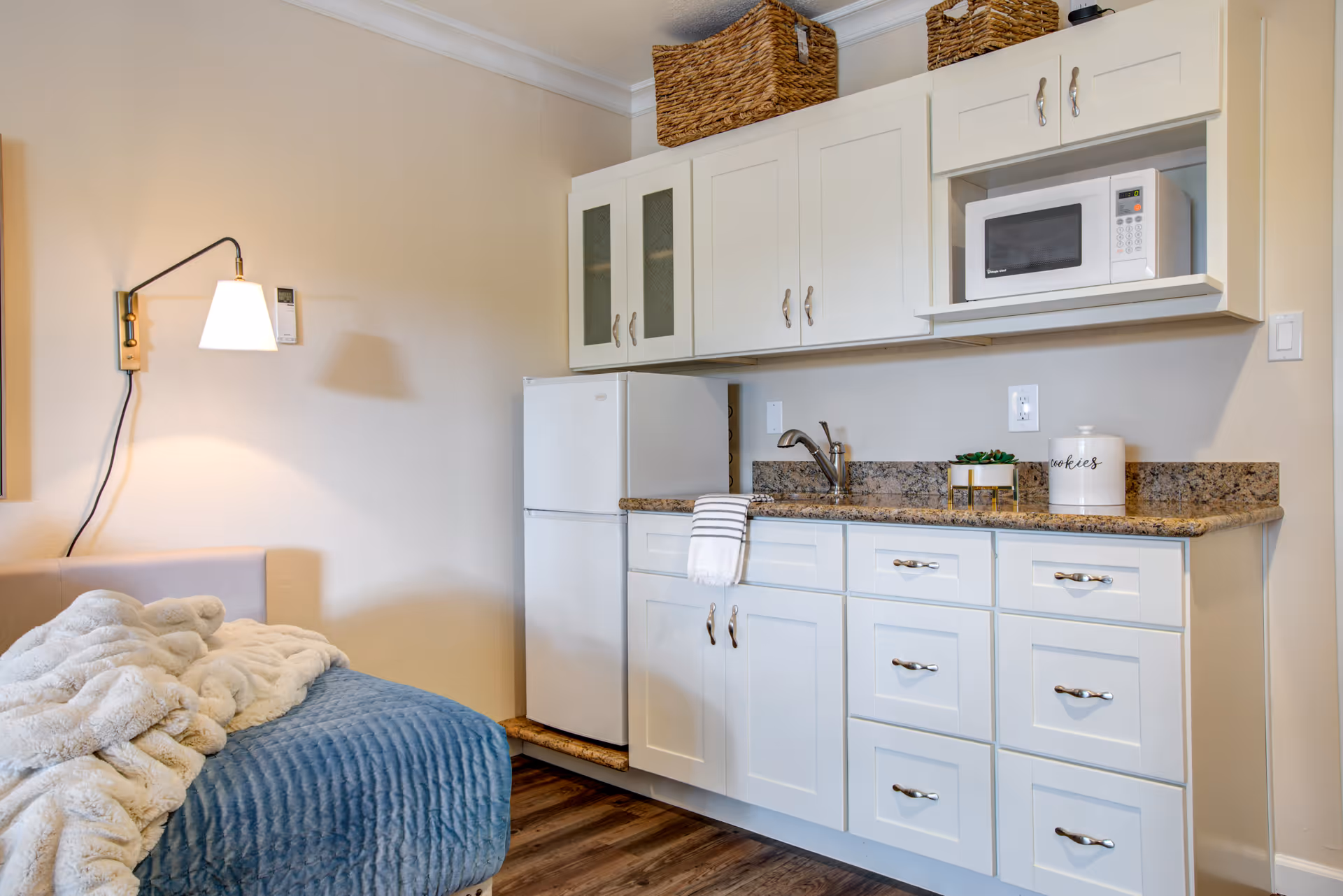 A small kitchenette area with white cabinets, a granite countertop, a white refrigerator, and a microwave. There is a sink with a faucet and a striped towel hanging on the cabinet handle. On the countertop, there is a small plant and a white container labeled 'cookies'. To the left, part of a bed or couch with a blue quilted cover and a white fluffy blanket is visible. A wall-mounted lamp with a white shade is above the bed or couch.