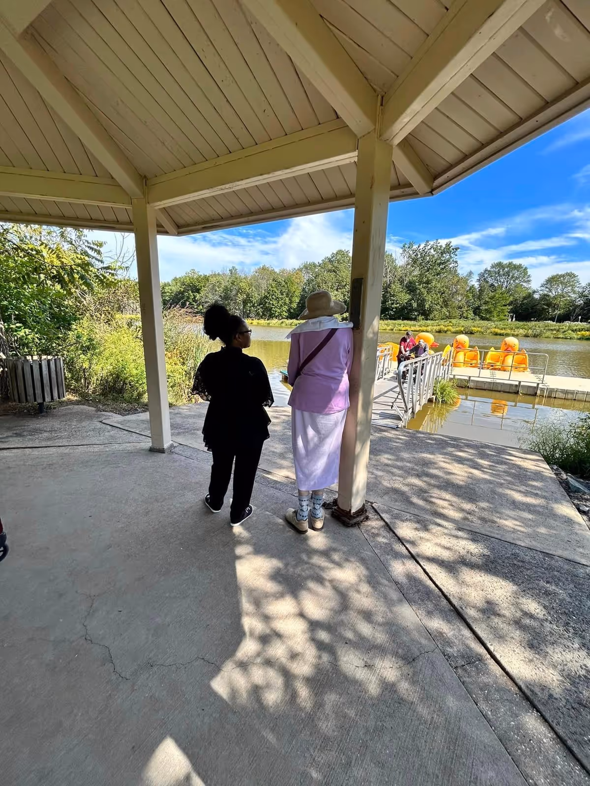 Two people standing under a covered pavilion near a lake. One person is wearing a black outfit and the other is wearing a light purple top, white skirt, and a wide-brimmed hat. In the background, there is a dock with several bright orange pedal boats on the water and trees surrounding the lake under a blue sky with some clouds.