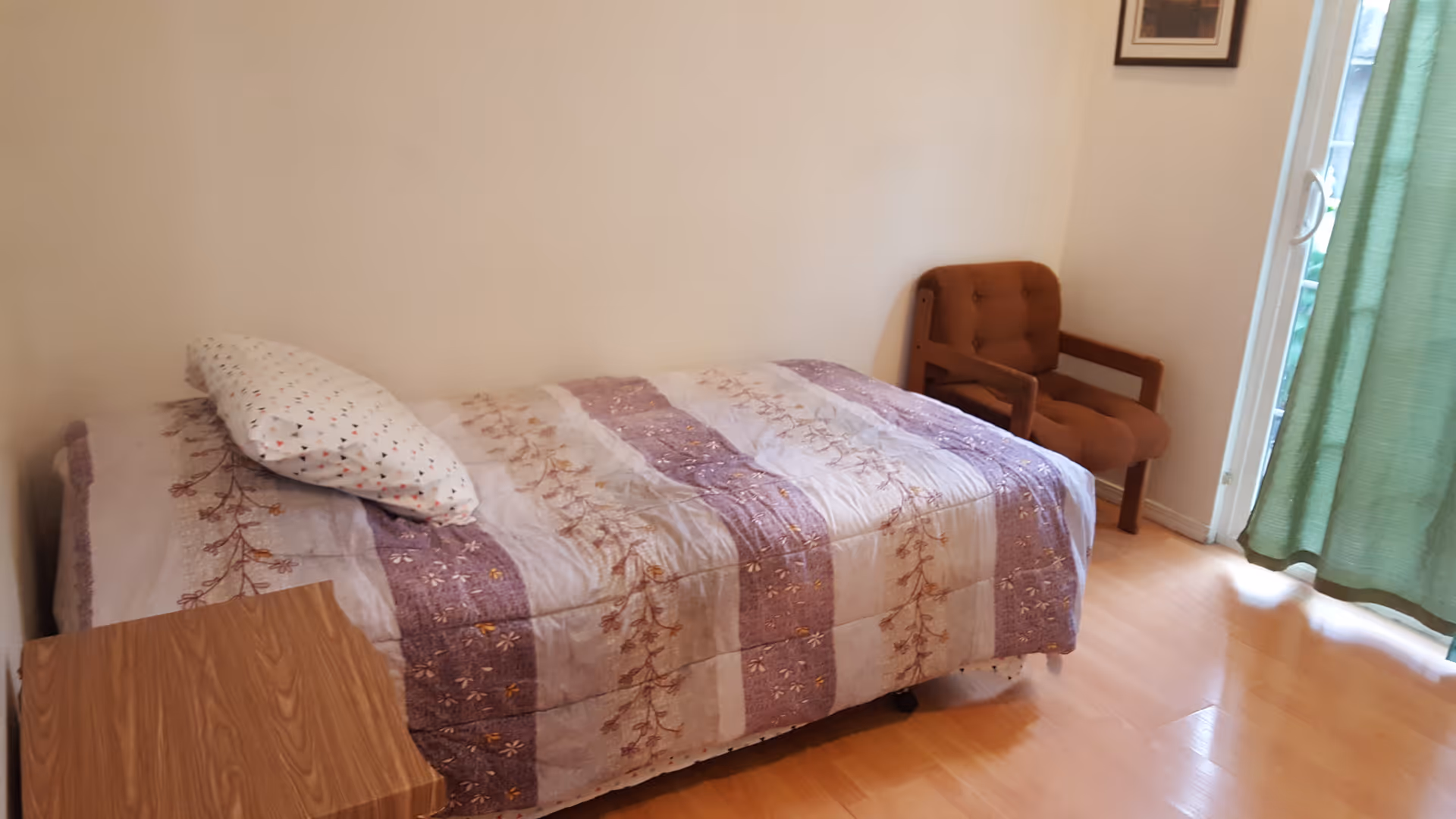 Small bedroom with a twin bed covered in a patterned quilt, a wooden nightstand, an upholstered chair, and a sliding glass door with green curtains.