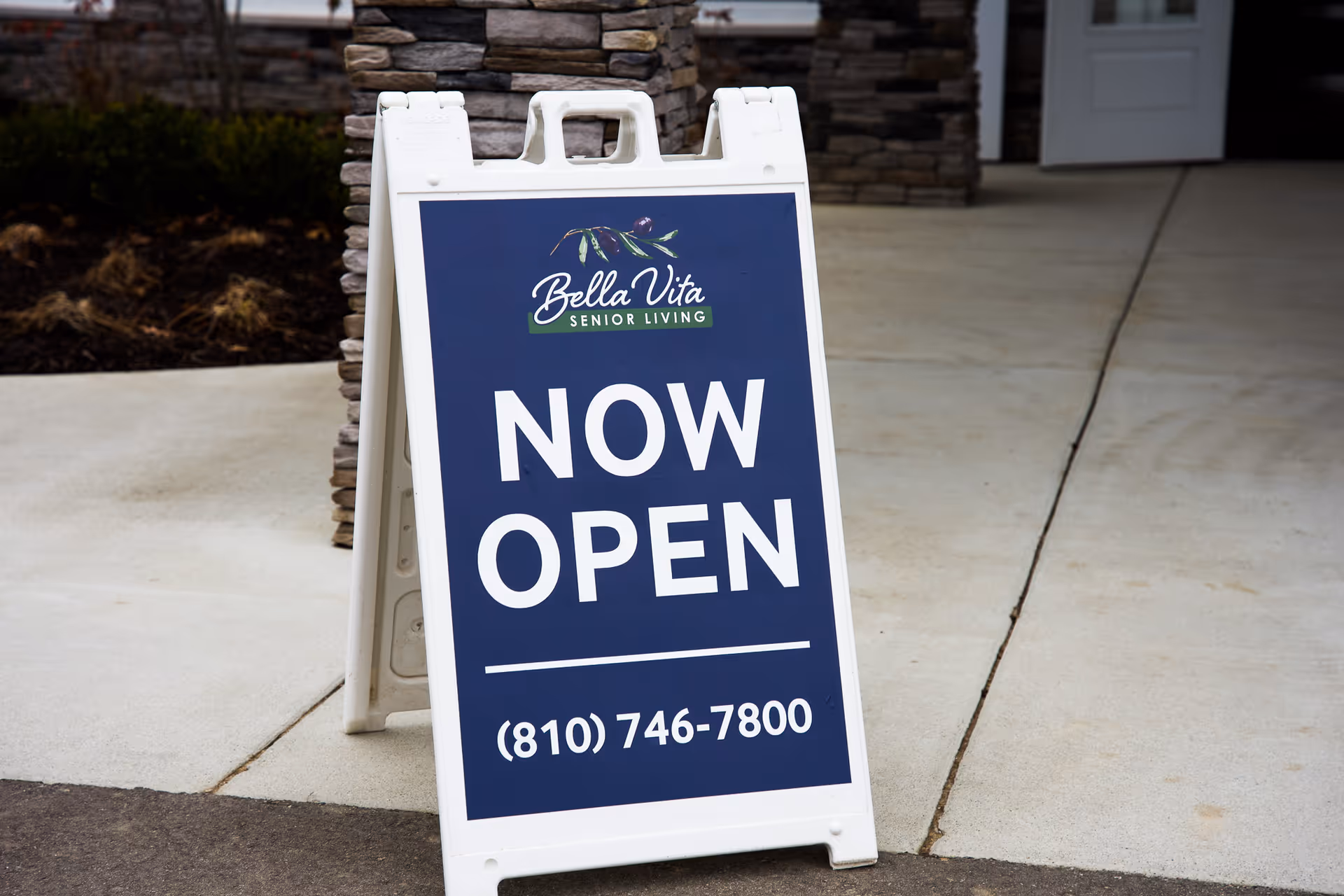 A sandwich-board sign reading "Bella Vita Senior Living NOW OPEN" stands on the sidewalk outside a facility entrance.