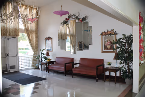 A waiting area with two maroon leather sofas and two wooden side tables with lamps and plants. The walls are decorated with framed floral paintings and mirrors. Large windows with gold curtains allow natural light to enter. The floor is tiled and there is a black mat near the entrance door.