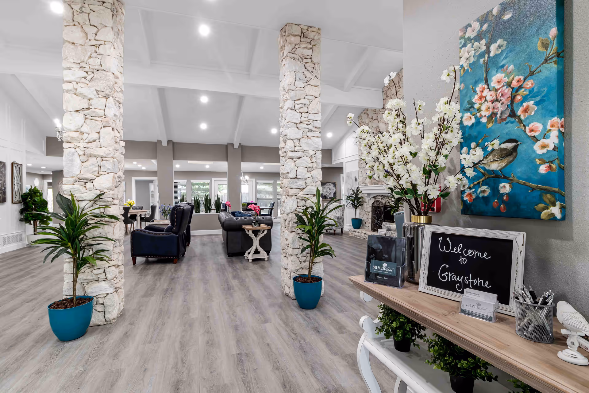 A spacious and well-lit living area with stone pillars, wooden flooring, and comfortable seating including armchairs and sofas. There are several potted plants and decorative flowers, a fireplace in the background, and a small table with a chalkboard sign that reads 'Welcome to Graystone' alongside brochures and a painting of a bird on a flowering branch on the wall.