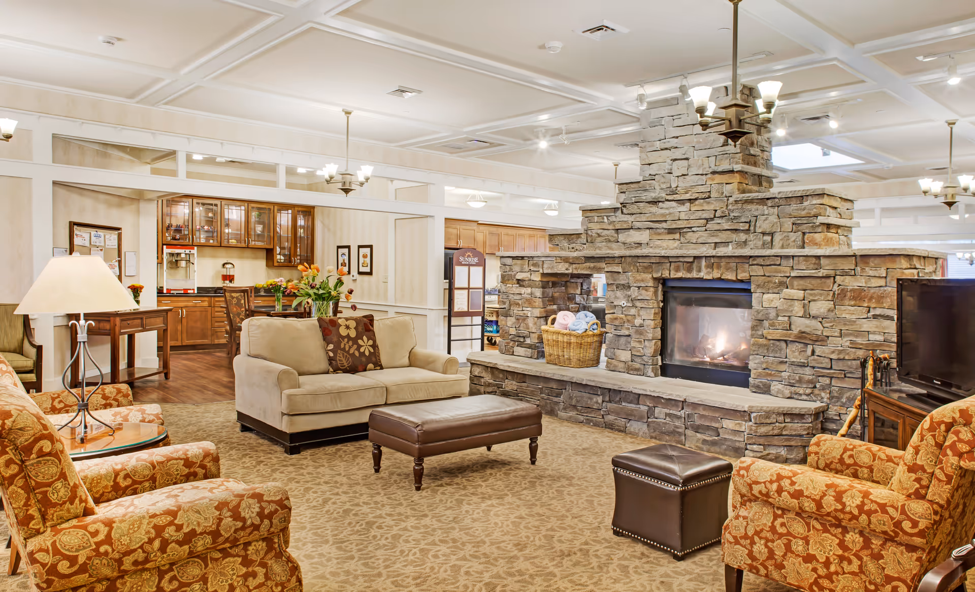 Spacious senior living lounge with sofas and armchairs arranged around a large stone fireplace.