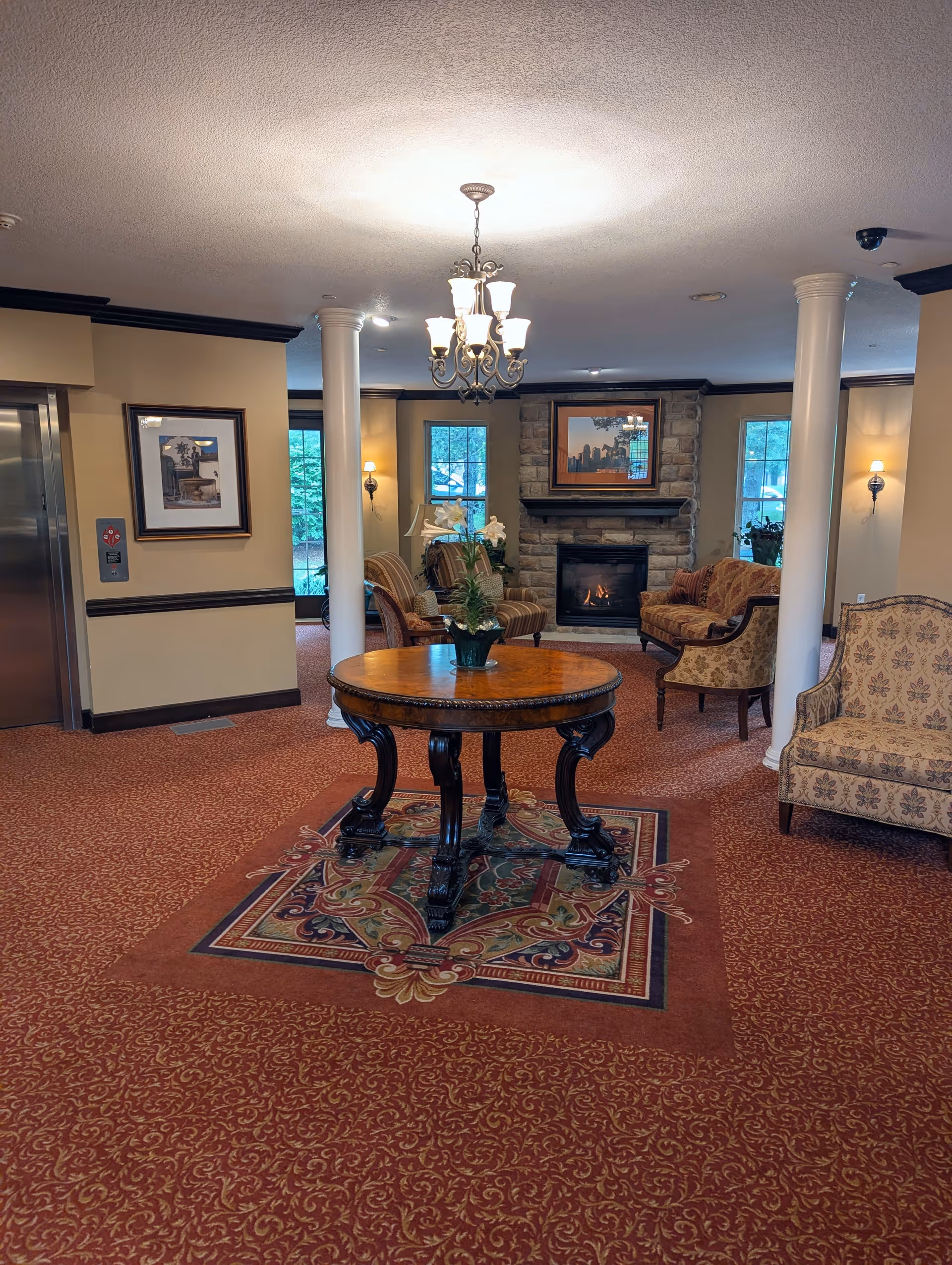A cozy senior living community lounge area with a round wooden table on an ornate rug in the center. The room features patterned carpet, two white columns, a stone fireplace with a framed picture above it, several upholstered chairs and sofas, wall sconces, and a chandelier hanging from the ceiling. There is an elevator on the left side and windows letting in natural light.