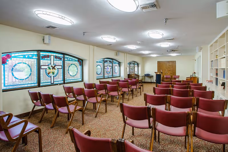A carpeted meeting room with rows of red chairs facing a podium and stained-glass windows along one wall.
