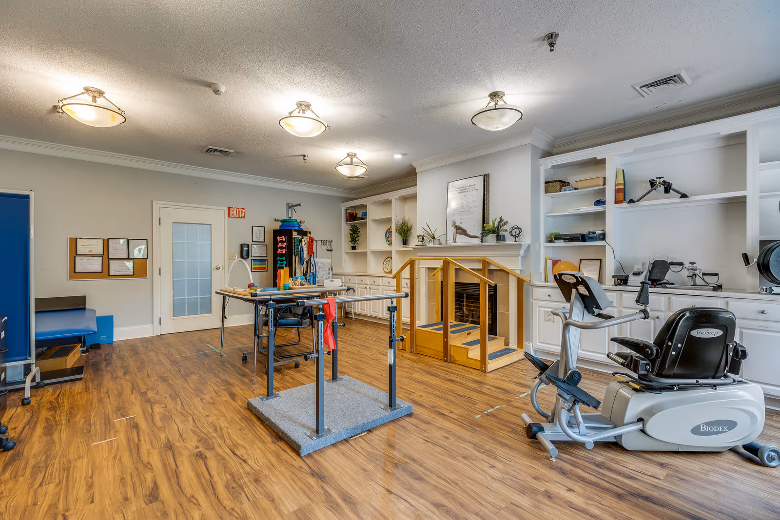 A rehabilitation or physical therapy room with wooden flooring, parallel bars for walking exercises, a stationary exercise bike, and various therapy equipment. The room has white built-in shelves with plants and equipment, a small set of stairs with handrails, and a door with a frosted glass panel. The lighting consists of ceiling-mounted fixtures.
