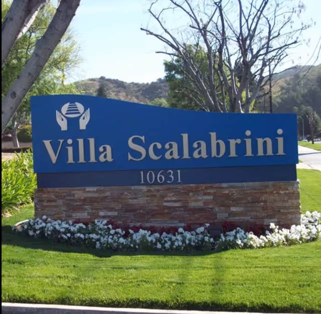 Outdoor sign for Villa Scalabrini Retirement Center with the address number 10631, surrounded by green grass, white flowers, and trees with hills in the background.
