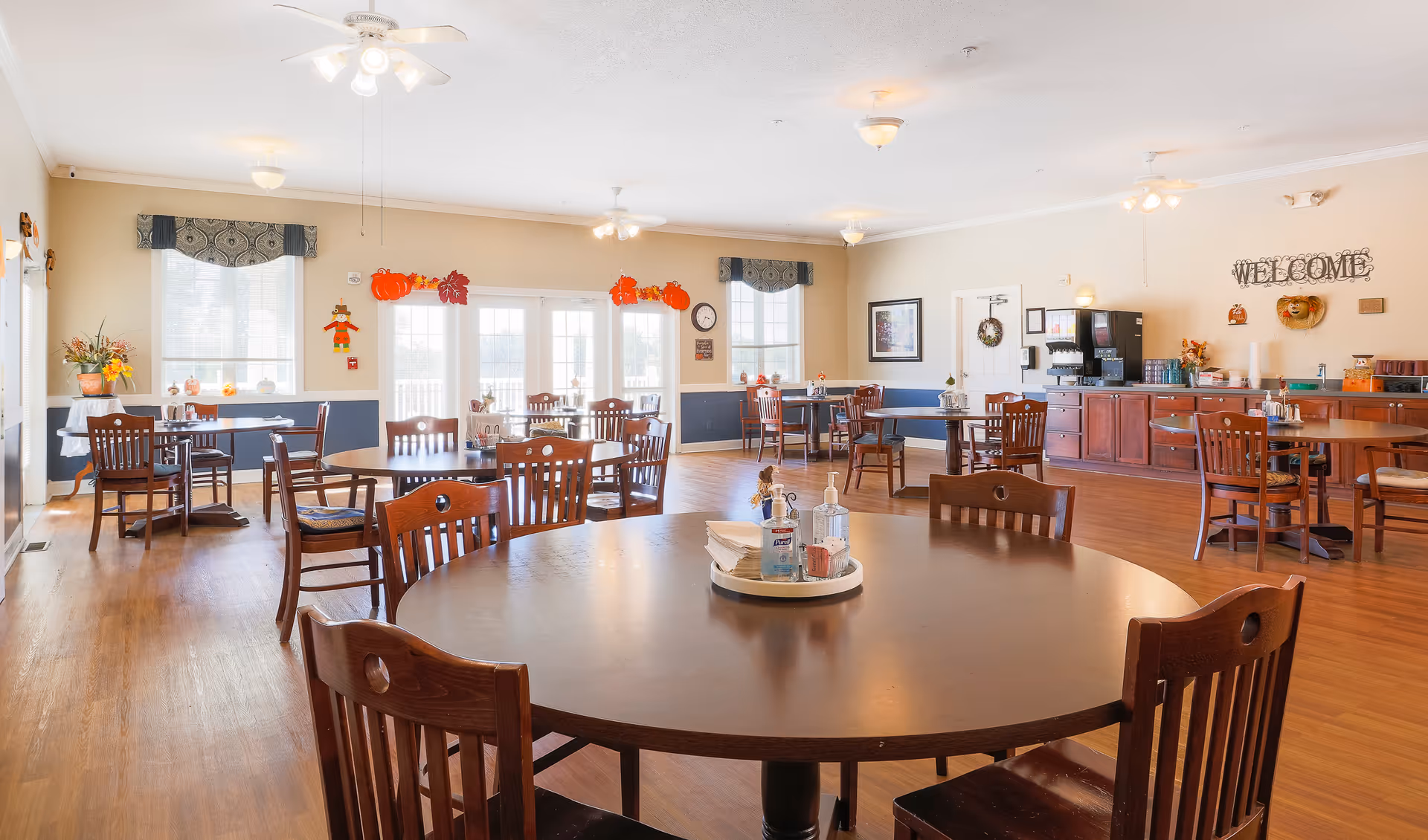 A bright and spacious dining room in an assisted living facility with multiple wooden tables and chairs arranged neatly. The room features large windows with valances, fall-themed decorations including pumpkins and scarecrows, and a buffet area with a coffee machine and various items. The walls are painted in light colors with a 'WELCOME' sign displayed.
