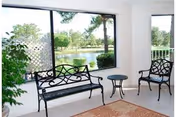 A bright indoor seating area with two black metal chairs and a matching bench around a small round table. Large windows provide a view of a green outdoor landscape with trees and a pond. A potted plant is visible on the left side and a patterned rug covers part of the floor.