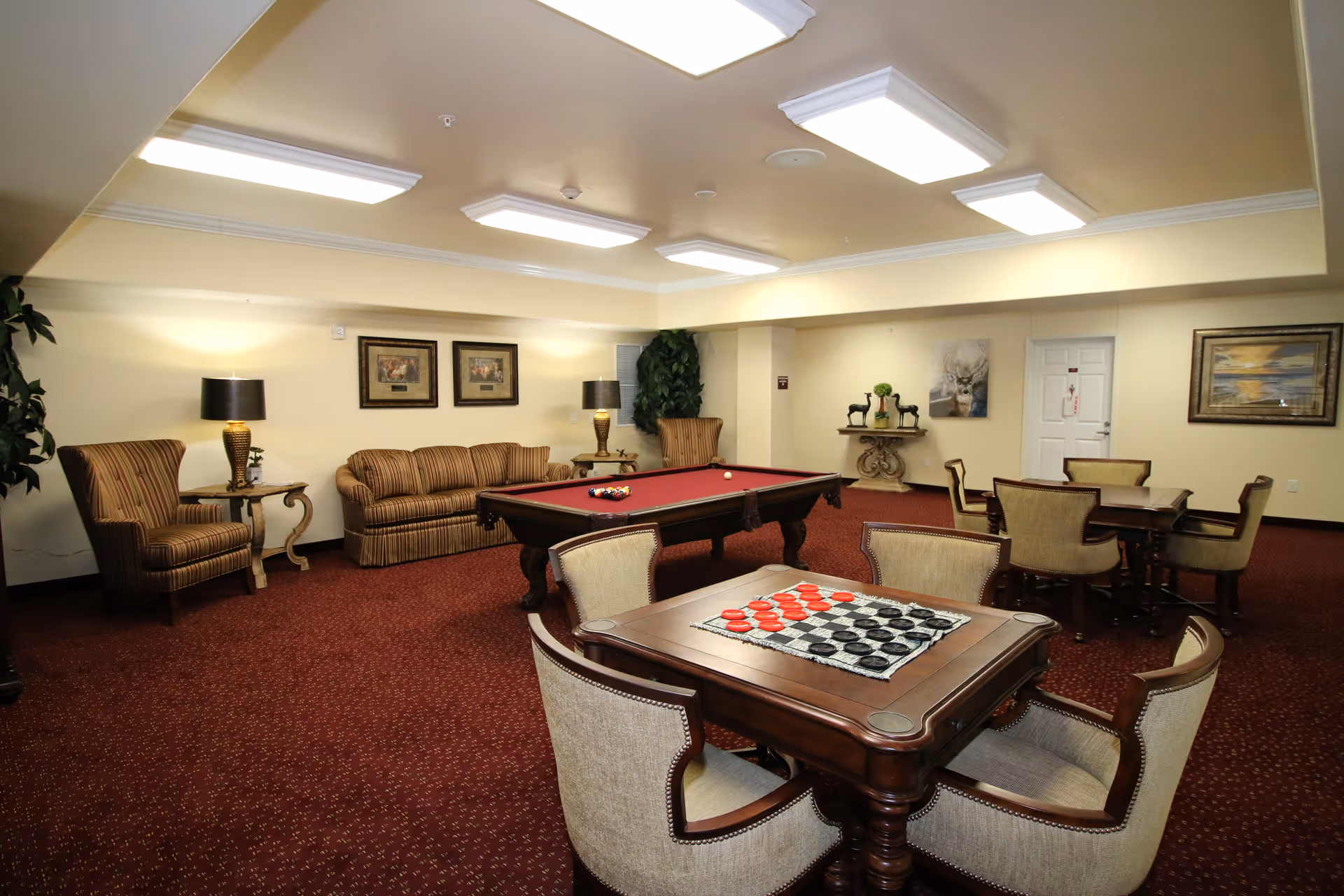 A spacious game room with a red pool table in the center, a table with a checkers board in the foreground, and several chairs and tables arranged around the room. There is a striped sofa and armchairs along the left wall, with two lamps on side tables. The walls are decorated with framed pictures and artwork, and the room has a warm, inviting atmosphere with carpeted flooring and bright ceiling lights.