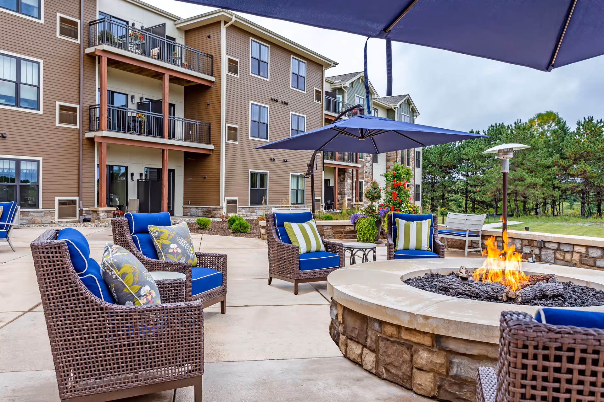 Outdoor patio with wicker seating, blue cushions, umbrellas and a central stone fire pit in front of a multi-story residential building.