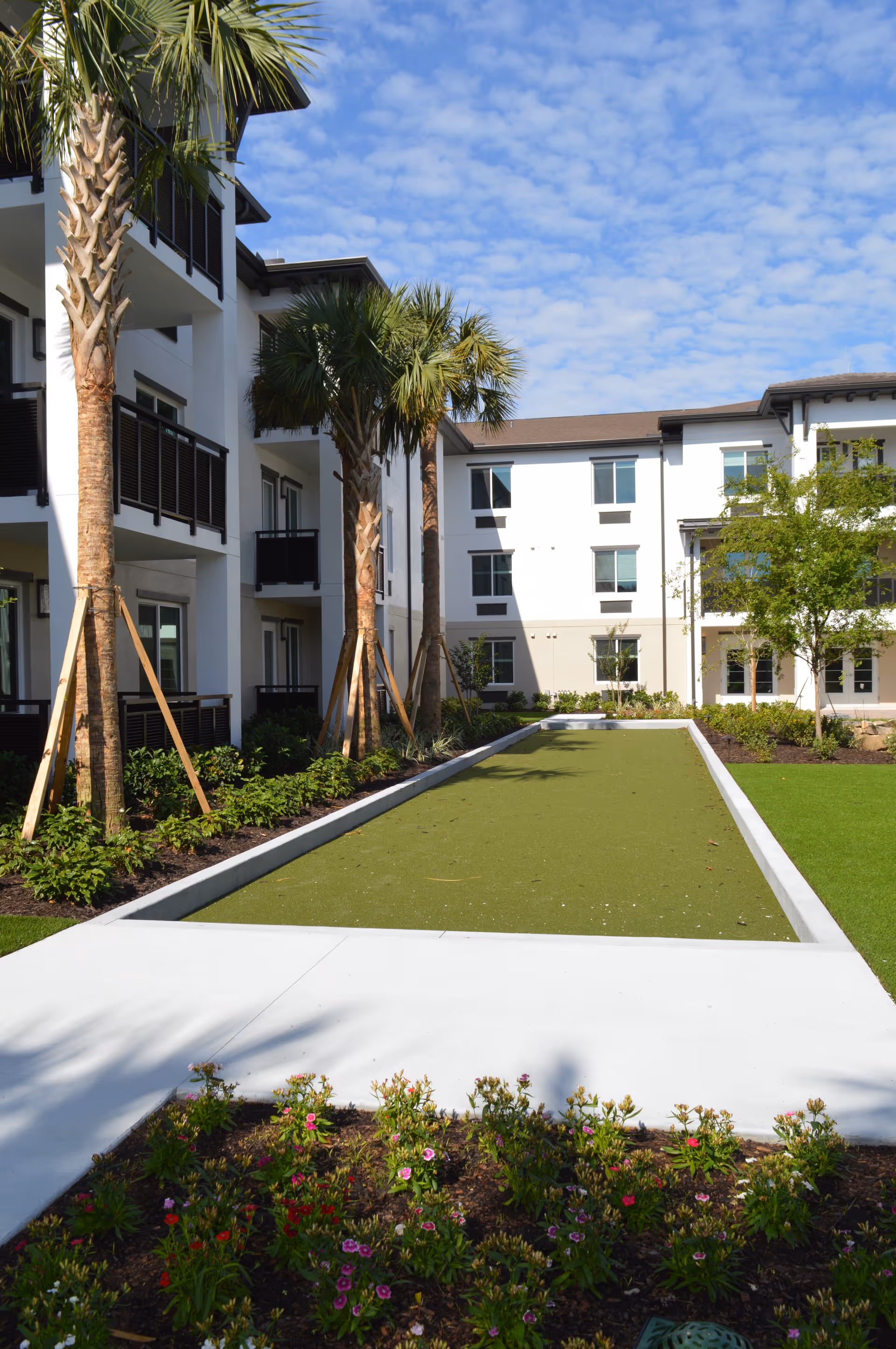 Outdoor courtyard at a senior living facility featuring a long rectangular green lawn or bocce court, palm trees, flowers, and surrounding three-story white buildings.