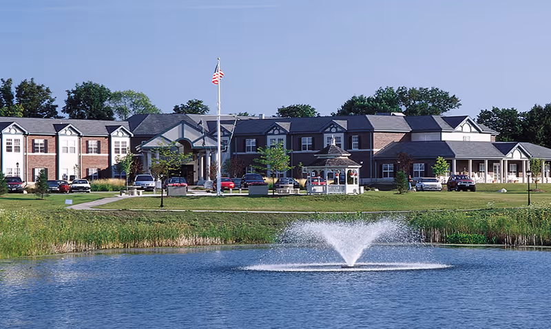 Front exterior of a brick senior living building with a flagpole, gazebo, parked cars, and a pond with a fountain in the foreground.