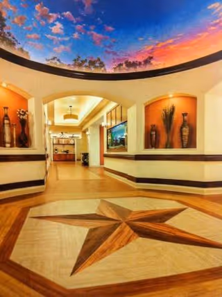 Interior hallway of a senior living facility with a large star design on the wooden floor, arched doorways, decorative vases in wall niches, and a ceiling mural depicting a colorful sky with clouds and trees.