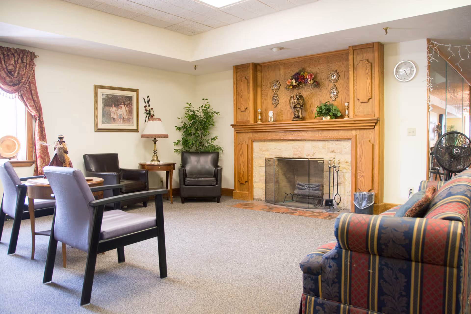 A cozy living room area in an assisted living facility featuring a stone fireplace with a wooden mantel decorated with figurines and plants. The room has a patterned sofa, two black armchairs, a small round table with two chairs, a side table with a lamp, a potted plant, a wall clock, and a window with red curtains.